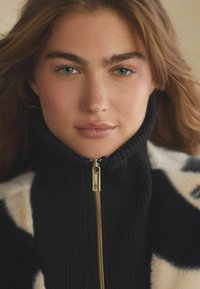 Mustast ribakraega sviiter, millel on kuldne tõmblukk ja pehme kreemja ja musta mustriga kate. Sile tekstuur, lähedal sobiv disain.