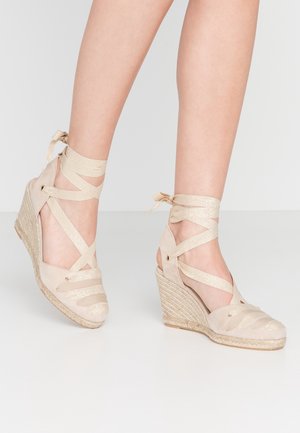 Beige kilehæl espadrillos med krydsende ankelbånd, båret på bare ben mod en ensfarvet baggrund.