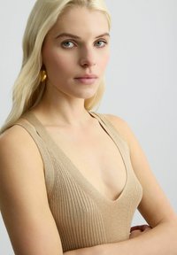 Top a coste beige con scollatura a V profonda e spalline larghe. Il modello indossa orecchini color oro. Sfondo grigio chiaro uniforme.