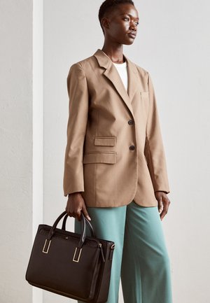 Femme portant un blazer beige et un pantalon bleu clair, tenant un sac à main noir avec des ferrures dorées, debout contre un mur blanc uni.