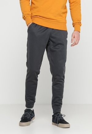 Man draagt donkergrijze joggerbroek, zwarte sneakers met beige zolen en een oranje sweatshirt, staand tegen een witte achtergrond.