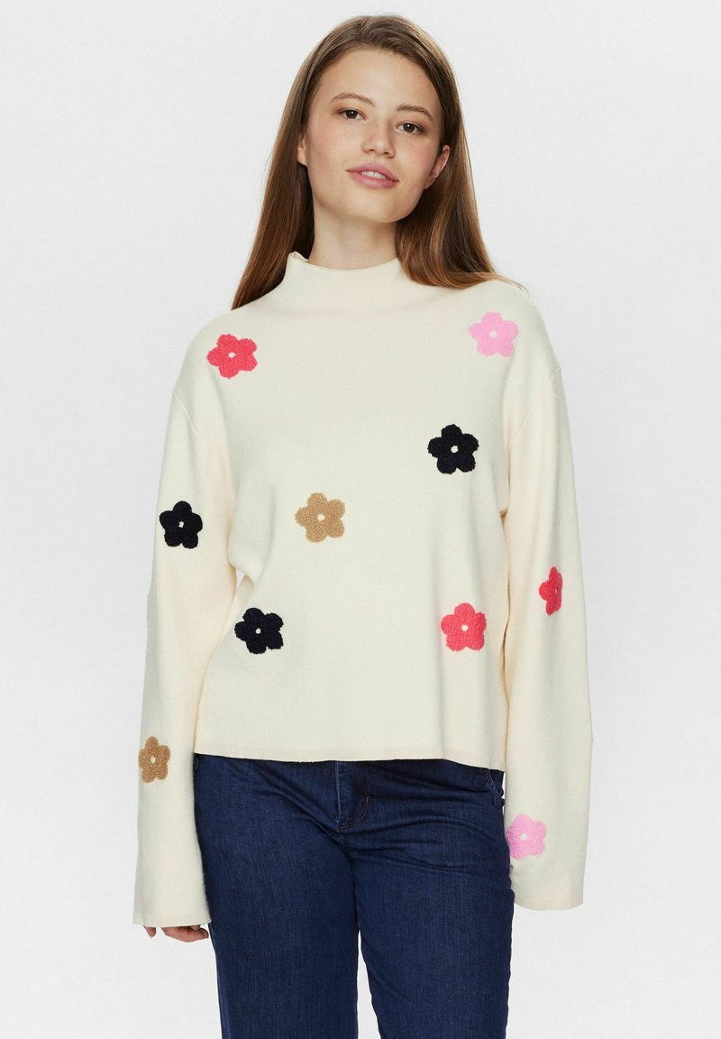 Cremfarbener Pullover mit hohem Kragen, verziert mit bunten Filzblumen-Applikationen in Pink, Schwarz und Braun, kombiniert mit dunklen Jeans.