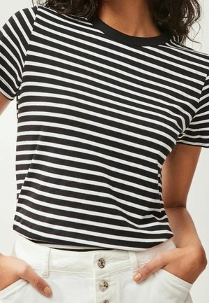 Chemise à manches courtes rayée horizontalement noir et blanc portée avec un pantalon blanc taille haute à boutons métalliques, mains dans les poches.