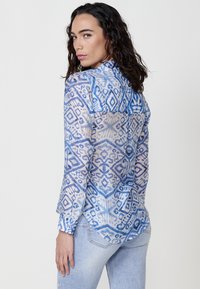 Camicia azzurra trasparente con bottoni, caratterizzata da un motivo geometrico, maniche lunghe e orlo curvo. Il tessuto ha una finitura leggera e testurizzata.