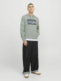 Felpa verde chiaro con logo frontale, tasca a marsupio, abbinata a jeans in denim scuro a vestibilità ampia e sneakers bianche con suola color beige chiaro.