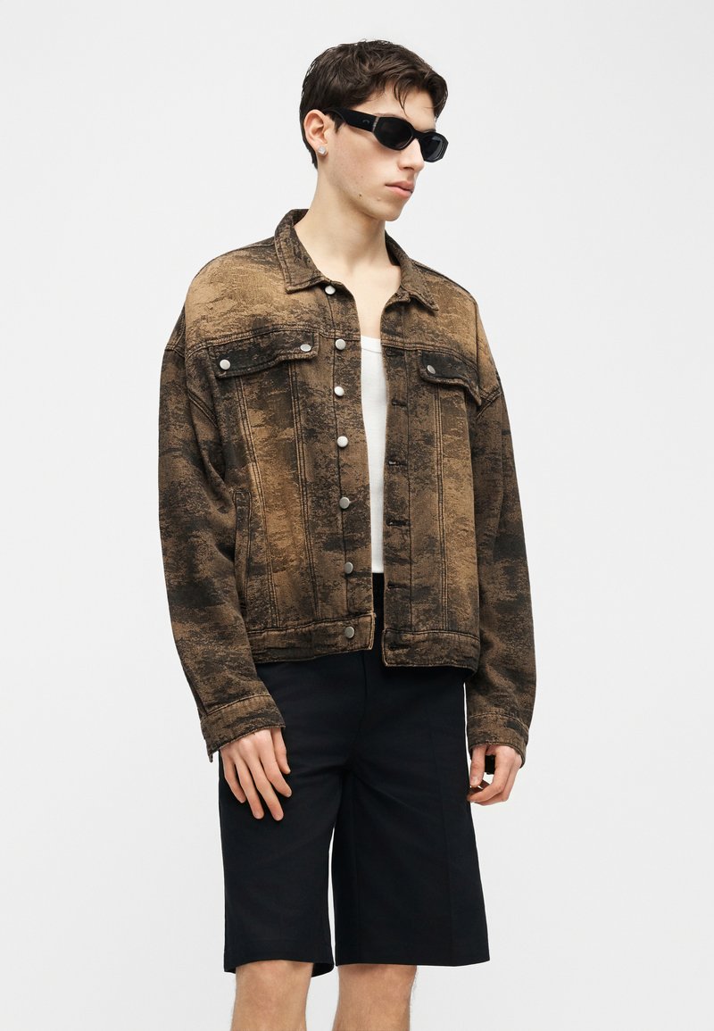 Giovane uomo che indossa occhiali da sole neri, giacca marrone testurizzata con bottoni, maglietta bianca e pantaloncini neri fino al ginocchio su sfondo semplice.