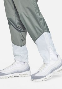 Calças leves corta-vento em verde-oliva e branco com fechos de correr nos punhos. Combinadas com ténis brancos e cinzentos com sola texturizada.