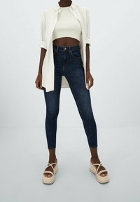 Camisa blanca de botones sobre un top cropped de canalé, combinada con jeans skinny azul oscuro y sandalias de plataforma beige con tiras entrelazadas.