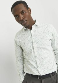 Camisa blanca de botones con un pequeño patrón floral azul; presenta un cuello clásico y mangas largas. Combinada con pantalones oscuros y un cinturón.