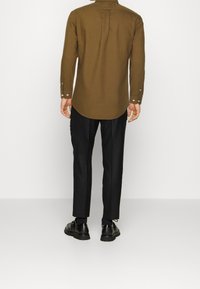 Camicia marrone a maniche lunghe con orlo curvo e polsini con bottoni, abbinata a pantaloni neri su misura e scarpe nere, vista da dietro.