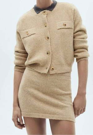 Beige strikket cardigan med knapper og to klaplommer, kombineret med matchende tætsiddende strikket mininederdel, båret med ærmer let skubbet op.