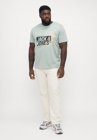 Light green cotton t-shirt featuring "JACK & JONES" graphic print in black and yellow. Paired with white pants and grey sneakers.