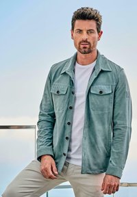 Hellgrüne, knöpfbare Jacke mit zwei Brusttaschen, einem Kragen und langen Ärmeln. Wird über einem weißen T-Shirt getragen und mit hellen Hosen kombiniert.