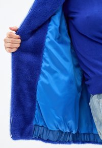 Giacca in pelliccia sintetica di un blu vivace, con una morbida fodera in satin, chiusura con zip e orlo arricciato. Mani tengono il bordo non chiuso per mostrare l'interno.