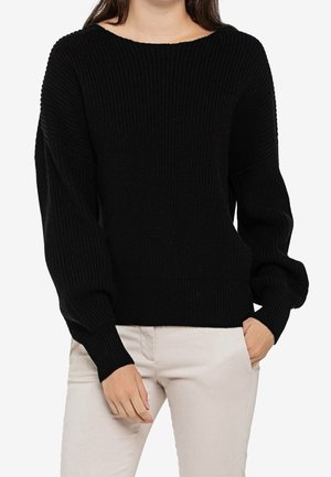Maglione nero lavorato a maglia con texture a costine, maniche ampie e scollatura arrotondata. Abbinato a pantaloni chiari, mani nelle tasche.