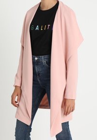 Cardigan rosa chiaro aperto sul davanti in un tessuto morbido, con maniche lunghe e una vestibilità rilassata, indossato sopra una maglietta nera con scritte colorate.