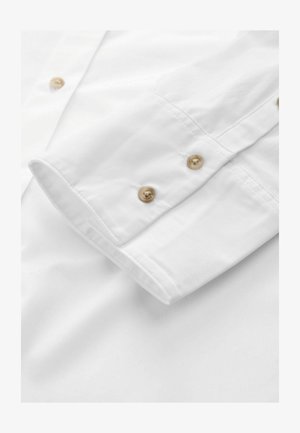 Poignet de chemise en coton blanc avec deux boutons beiges, couture propre, et texture lisse, partiellement retroussé sur fond blanc.