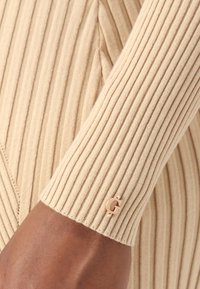 Manica a costine beige con bordo arrotondato; presenta un piccolo accento con logo dorato. Texture liscia, design aderente.
