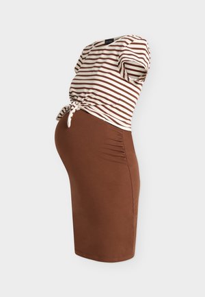 Zwangere vrouw staat, draagt een gestreept kortemouwtopje dat in de taille is geknoopt, een bruine rok, zwarte open-toe mule schoenen en draagt een zwarte tas.