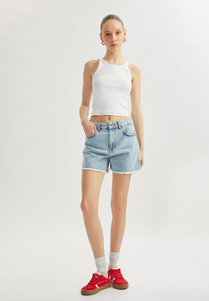 Top sin mangas blanco de canalé y pantalones cortos de mezclilla azul claro con bordes deshilachados. Zapatillas rojas con detalles blancos y calcetines grises completan el atuendo.