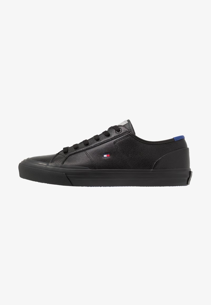 Basket basse en cuir noir avec lacets noirs, semelle en caoutchouc plate, petit logo rouge, blanc et bleu sur le côté et languette bleue au talon.