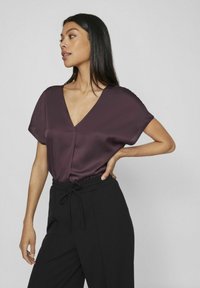 Tamsiai raudonas satino palaidinės, su V formos iškirpte, trumpomis rankovėmis ir centrinėmis siūlėmis, derinamas su juodomis kelnėmis su aukštu liemeniu ir raišteliu.