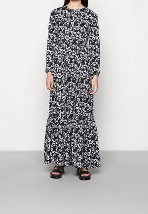 Vrouw draagt een zwarte maxi-jurk met lange mouwen en witte bloemmotieven en zwarte sandaal met open tenen, staand tegen een effen lichte achtergrond.