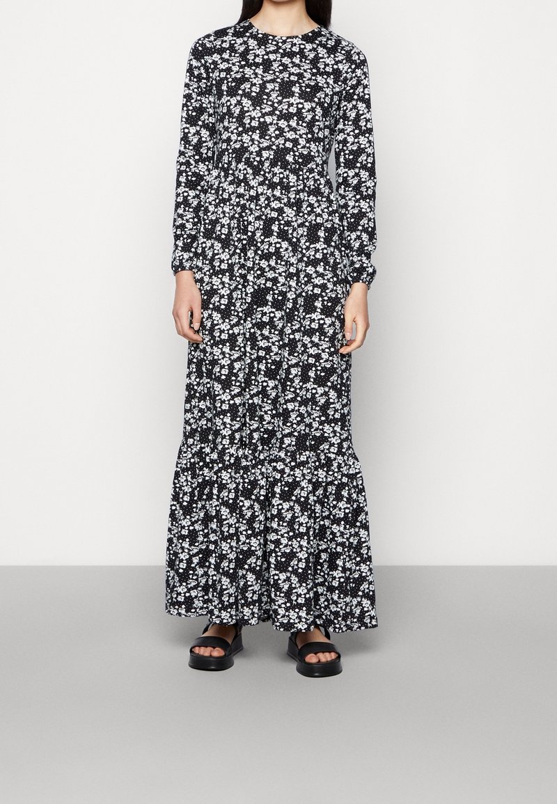 Femme portant une robe maxi noire à manches longues avec un motif floral blanc et des sandales noires à bout ouvert, debout devant un fond clair uni.