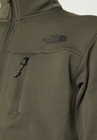 Olivově zelená fleecová bunda na zip s logem na hrudi, hladkou texturou, ochranou brady a kapsou na zip na levé straně.