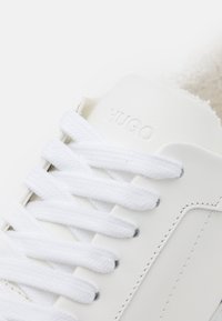Białe skórzane sneakersy z teksturowaną powierzchnią, z płaskimi białymi sznurówkami i subtelnymi szarymi akcentami. Wytłoczone logo "HUGO" na języku.