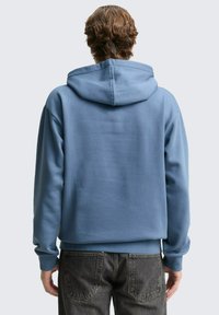 Zils kapuces džemperis ar plašu kapuci, mīkstu tekstūru, ribotas aproces un apakšmalu, kombinēts ar tumši pelēkiem džinsiem ar aizmugurējām kabatām.