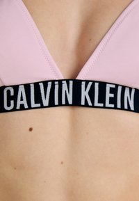 Rosa Sport-BH mit tiefem V-Ausschnitt, ausgestattet mit einem schwarzen elastischen Band, auf dem "CALVIN KLEIN" in Weiß gedruckt ist. Glatte Textur, minimales Design.