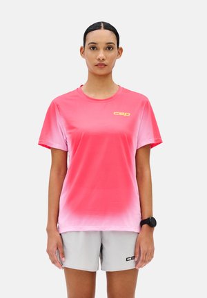 Femme aux cheveux foncés portant un t-shirt de sport à dégradé rose et un short gris clair, avec une smartwatch noire, face à l'avant sur un fond uni.