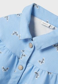 Camicia in denim blu chiaro con collo a cravatta, motivi a farfalla e design arricciato. Presenta dettagli con bottoni in metallo per la chiusura.