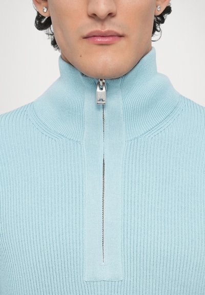 Pull à fermeture éclair bleu clair avec texture côtelée et col montant. Dispose d'une fermeture éclair argentée avec un tirette logo.