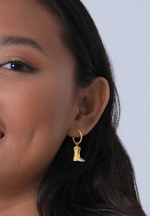 Boucles d'oreilles en or en forme de créoles, ornées de charm Bracelet en forme de petite botte de cowboy avec un relief et un design détaillés. L'arrière-plan est simple et discret.