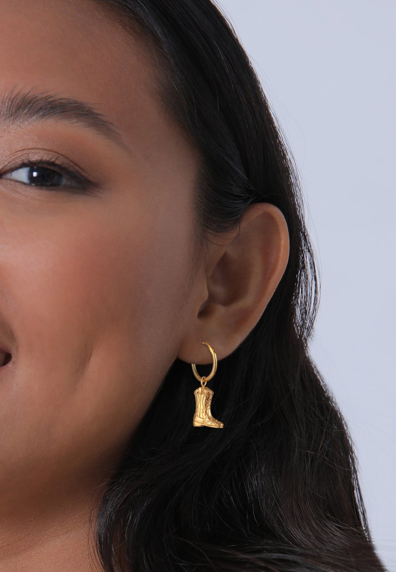 Boucles d'oreilles en or en forme de créoles, ornées de charm Bracelet en forme de petite botte de cowboy avec un relief et un design détaillés. L'arrière-plan est simple et discret.