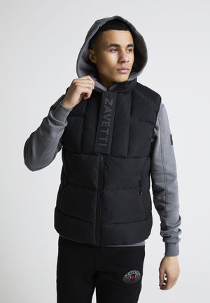 Ung mann iført en svart polstret vest med "ZAVETTI"-stropp over en grå hettegenser, justerer vestens krage, og ser mot høyre.