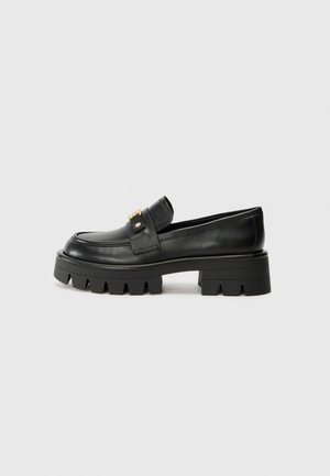 Schwarzer Leder-Slipper mit grober, profilierten Sohle und goldener Schnallenverzierung an der Seite, seitlich auf hellem Grauhintergrund gezeigt.