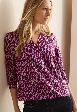 Blusa a fantasia animale viola con scollo a V, maniche lunghe e vestibilità rilassata. Il design presenta motivi bianchi irregolari su uno sfondo scuro.