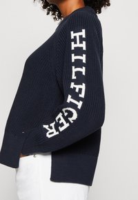 Marineblauer Strickpullover mit geripptem Textur und weißem "HILFIGER" Schriftzug auf dem Ärmel. Bietet eine lockere Passform und Seitenschlitze für mehr Bewegungsfreiheit.