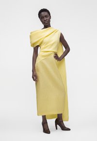 Femme portant une robe midi jaune texturée à une épaule avec un détail drapé et des chaussures marron à talons hauts, debout la main sur la hanche.