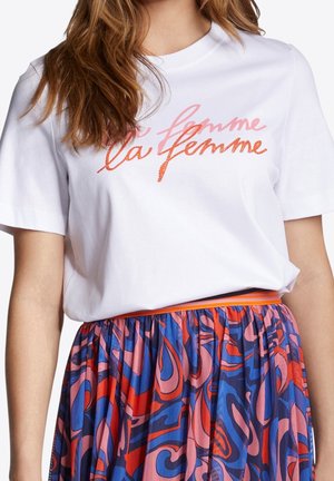 Kobieta w białym T-shircie z napisem "la femme" w różowo-pomarańczowym stylu pisma, zestawionym z wielokolorową spódnicą o abstrakcyjnym wzorze.