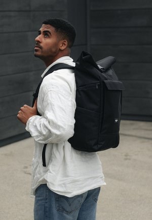 Black roll-top backpack with zippered pocket, adjustable straps, and fabric texture; worn over a white long-sleeve shirt and denim jeans.