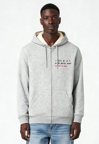 Grijze ritssluiting hoodie met een zachte, crème-kleurige fleece voering. Voorzien van voorzakken en geborduurde tekst in het rood en zwart op de borst.