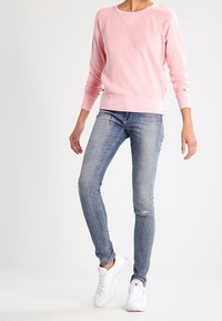 Hellrosa Pullover mit Rundhalsausschnitt und geripptem Saum, kombiniert mit ausgewaschenen blauen Skinny-Jeans und weißen Turnschuhen.