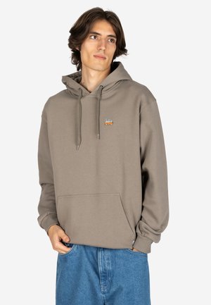 Young man wearing taupe hoodie with orange van embroidery on chest and blue jeans, hands partially in pockets, looking right.