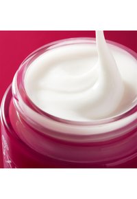 Witte crème in een doorzichtige pot met een roze deksel. De crème heeft een gladde, dikke textuur en is licht gedraaid terwijl het uit de pot lijkt te pieken.