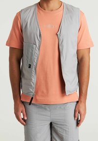 Gilet grigio senza maniche con zip frontale e tasca laterale, indossato sopra una maglietta color pesca con scollo rotondo. Tessuto liscio con una leggera texture.