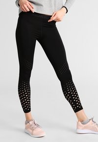 Osoba oblečená v černých legínách s perforovanými vzory a světle růžových teniskách se šedými ponožkami na jednobarevném pozadí.